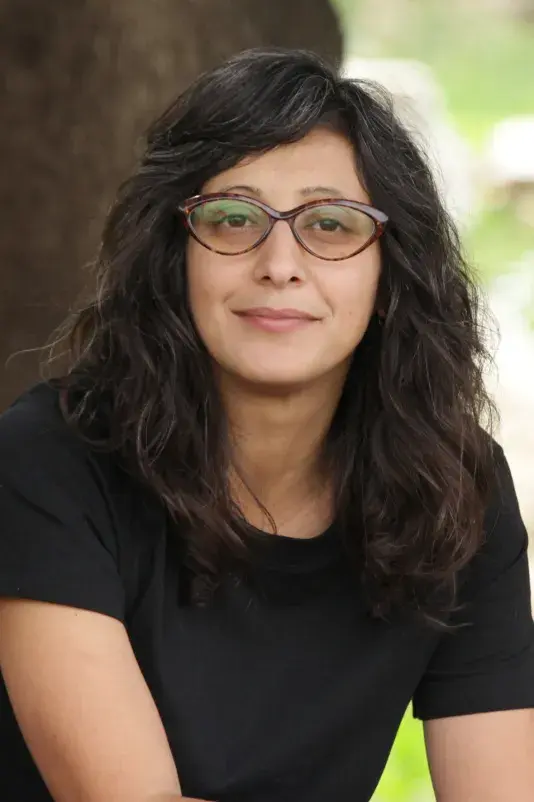 A portrait of Sahar Qawasmi, a woman with a medium-light skin tone and long wavy brown hair. She wears brown cat-eye glasses and a black t-shirt.