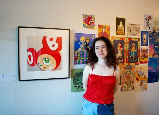 Woman with short dark hair wearing a bright red tank top is standing in front of a white wall with rows of bright posters. To the left of the woman there is a print of a red graphic figure with a black frame.