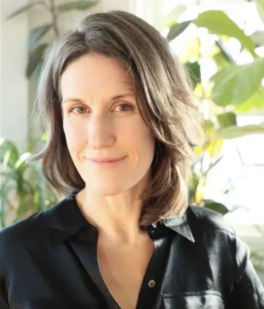 Portrait of a woman with dark hair in a black shirt standing in front of sunlight green leaves.
