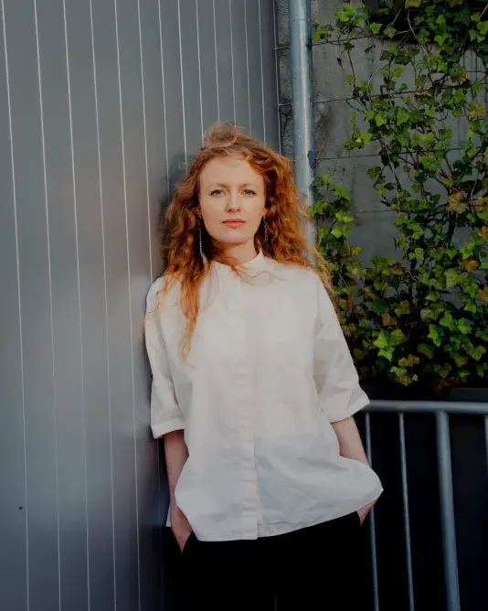 Portrait of Stefanie Hessler, a woman with a light skin tone and long red hair standing seriously in a white buttoned shirt with hands in pockets.