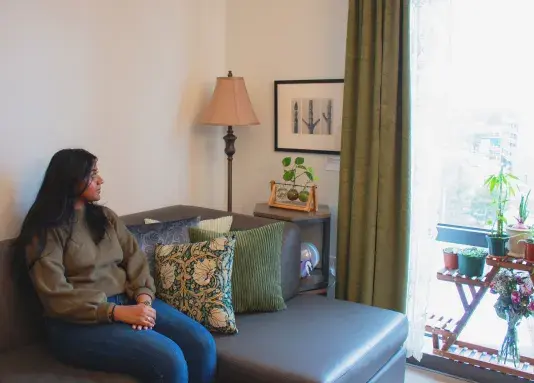 A woman sitting on a blue couch with three patterned pillows looking towards a window at the right side of the frame. There are plants at the base of the window. A black and white photograph in a black frame is to the left of the window, next to a standing lamp.