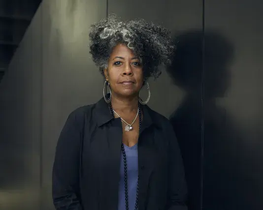 Portrait of Torkwase Dyson, a woman with a dark skin tone, short black and grey hair, and a soft smile, set against a black background.