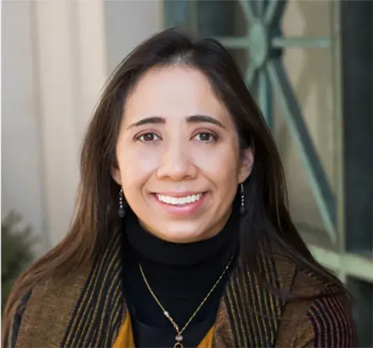 Portrait of Janelle Knox-Hayes, a woman with a medium-light skin tone, long brunette hair, and a broad smile wearing a yellow striped blazer.