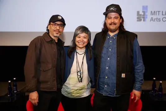 Three people standing on a stage in front of a projector screen. The two people on either side and taller than the person in the middle and are both wearing baseball hats.