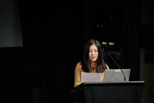 A woman with long dark hair is reading from a stack of paper while behind a podium with a tall microphone and a silver Mac laptop. The background is dark behind her except for two green squares.