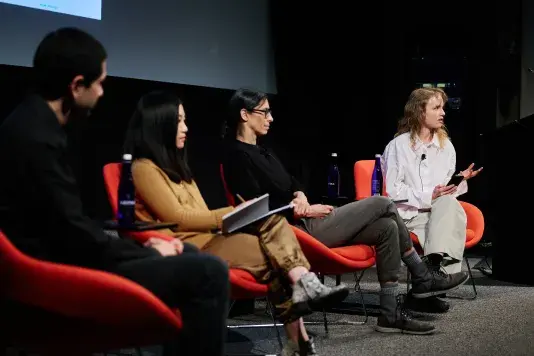 Four people sitting in a line in bright red chairs on a stage in front of a projector screen. The person furthest from the camera is speaking to the audience.