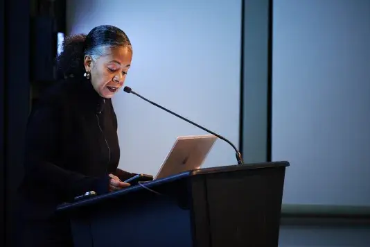 A woman is standing in a dimly lit room, behind a podium, and speaking into a microphone.