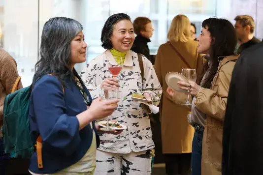 A group of people holding glasses of wine and plates of food gathered in an atrium lobby discussing with one another.