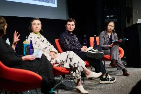 Four people sitting in a line in bright red chairs on a stage in front of a projector screen. The person closest to the camera is speaking to the panel.