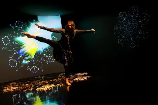 Still of a performance showcasing a woman spotlighted on stage with her arms out to both sides facing the camera and her right leg raised to hip height with foot pointed. A brightly colored geometric pattern in the background is juxtaposed by a black wall.