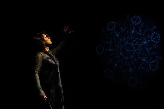 Still of a performance showcasing a woman spotlighted on stage with her left arm raised above her head. A faint colored geometric pattern in the background is juxtaposed by a black wall.