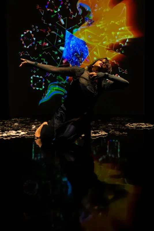Still of a performance showcasing a woman spotlighted on stage with her arms raised out to the right side of her body facing the camera. A brightly colored geometric pattern in the background is juxtaposed by a black wall.