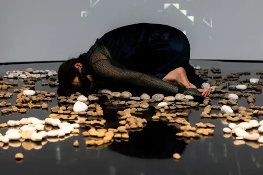 Still of a performance showcasing a woman curled up in fetal position spotlighted on stage laying amongst a collection of stones laid out in a pattern on a mirrored black floor.