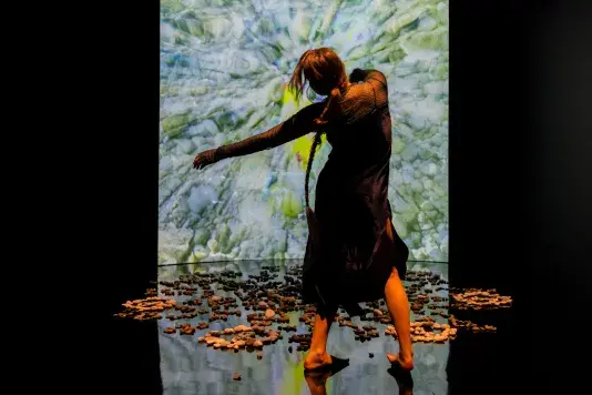 Still of a performance showcasing a woman spotlighted on stage with her arms out to the left side. A brightly colored geometric pattern in the background is juxtaposed by a black wall.