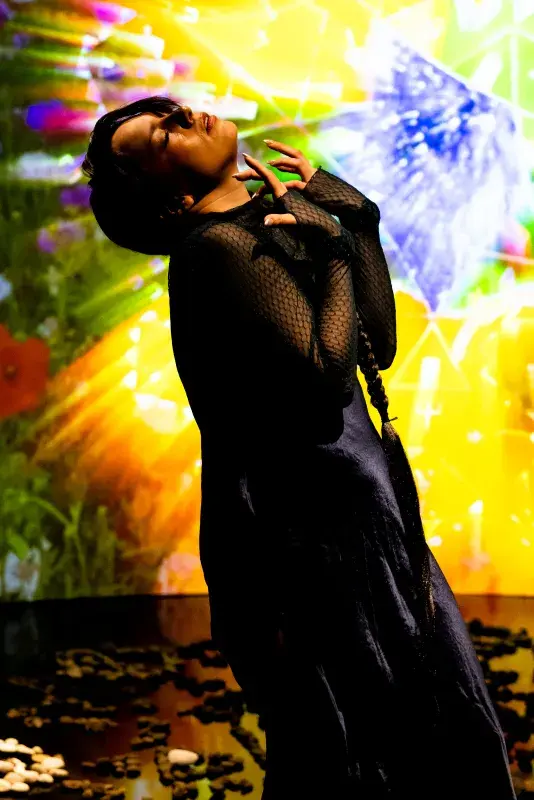 Still of a performance showcasing a woman spotlighted on stage with her head tilted up, arms raised in front of her chest. A brightly colored geometric pattern in the background is juxtaposed by a black mirrored floor.
