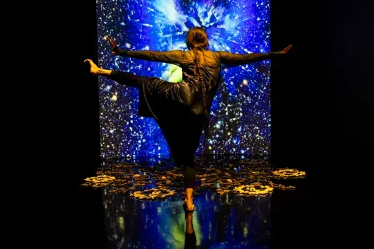 Still of a performance showcasing a woman spotlighted on stage with her arms out to both sides and her left leg raised to hip height with foot pointed. A brightly colored geometric pattern in the background is juxtaposed by a black wall.