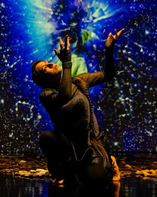 Still of a performance showcasing a woman spotlighted on stage with her head tilted up, arms raised and palms facing up. A brightly colored geometric pattern in the background is juxtaposed by a black wall.