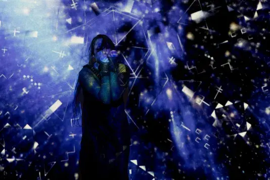 Still of a performance showcasing a woman spotlighted on stage with her arms raised in fists in front of her face. A brightly colored geometric pattern in the background is juxtaposed by a black wall.