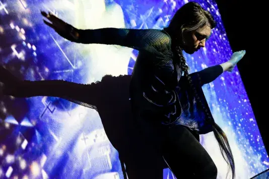 Still of a performance showcasing a woman facing the camera spotlighted on stage with her arms out to both sides and her right leg raised to hip height with foot pointed. A brightly colored geometric pattern in the background.