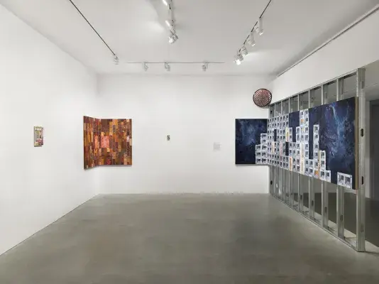 A blue cinder block-patterned painting wraps from scaffolding to the wall; a painted mirror and three other paintings hang throughout the gallery.