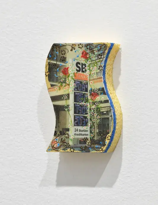 One side of a yellow and blue kitchen sponge is painted or printed with a view of a gas station. This scene is adorned with delicately painted flowers and other ornamental gestures.