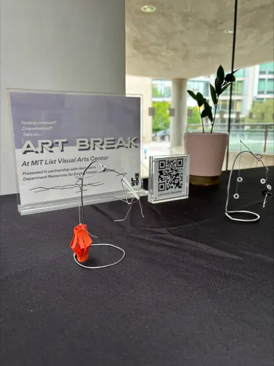 Plastic sign holder, a QR code to the right of the sign and a plant to the right of that on a table with a black tablecloth. The text reads "Art Break At MIT List Visual Arts Center"