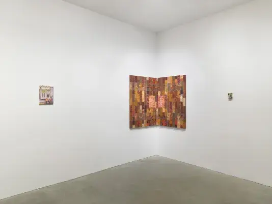 A corner of a white-walled gallery where three paintings are on display. On the left is a small painting of a street scene. In the middle, a painting featuring a brick-like pattern in shades of red, brown, pink, and orange is positioned in the corner of the room. On the right, a small painting on a sponge is mounted on the wall.