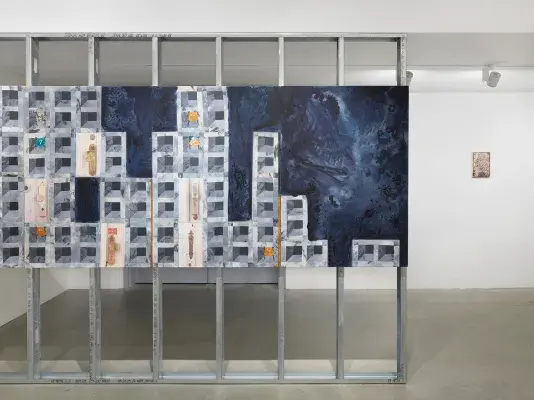A large blue painting hangs across steel beams. The painting features a cinder block-like motif, with scattered patches depicting doorknobs. In the background on the right, a smaller painting hangs on a white wall.