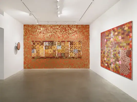 A tile-like painting hangs on an orange wall decorated with gold spray-painted motifs. To its left, a painted security mirror is mounted on a white wall. On the right wall, a painting featuring a brick pattern with flowers and collage elements is displayed.