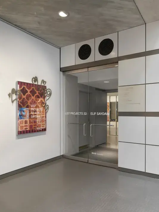An entryway to a gallery with glass doors. Text reading “List Projects 32: Elif Saydam” is displayed across the two doors. To the left of the entryway, a poster bearing the exhibition title hangs. The poster features a detail of a tile-like painting with gold spray-painted heart motifs that extend onto the adjacent white wall.