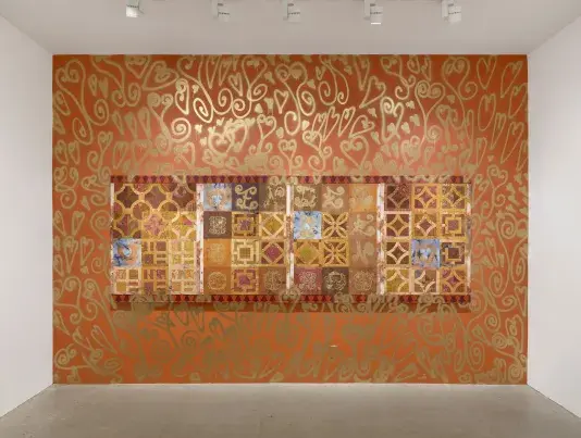 A painting with geometric breezeblock patterns in various shades of red and brown hangs on a rust-colored wall. Gold spray paint in exuberant heart shapes covers the surrounding wall, bleeding onto the wall and onto the canvas itself.