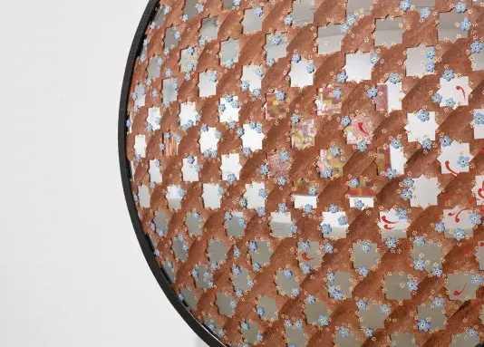 A close-up detail of a round convex security mirror that is covered by an interlocking brown painted star pattern. The uncovered “stars” are the exposed bare mirror reflecting the gallery space. Dispersed amongst the star pattern are small blue and yellow flowers with red brushtrokes scattered across the mirror.