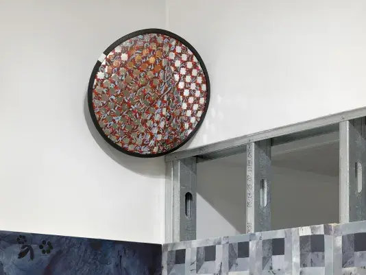 A close-up detail of a gallery corner. Above exposed steel beams hangs a convex security mirror painted with red and blue brushstrokes that form a star-shaped pattern of exposed mirror. The mirror reflects the gallery space. At the bottom of the image, the edges of a painting hung on both sides of the corner peek into the frame.