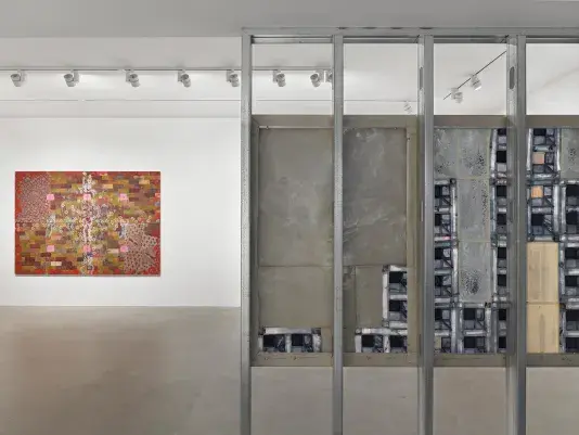 A view of a gallery space with two paintings on display. In the foreground on the right, the back side of a painting hangs across four steel beams, composed of blue patchwork pieces. In the background on the left, a painting featuring a brick pattern and flowers hangs on a white wall.