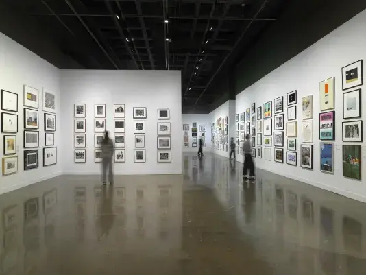 Installation view with framed artworks hanging on the walls salon style and four people standing looking at the art on the walls throughout the room.