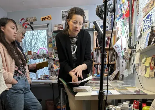 Woman bending over a table covered with art supplies pointing at a personal work of art. Two woman are standing to the left listening to the central figure speak.