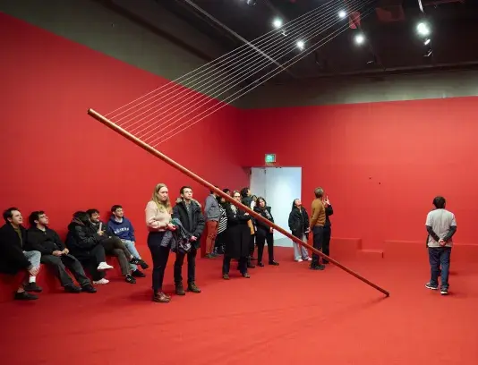 A completely red room is filled with people. There is a large metal pip suspended at an angle in the middle of the room.