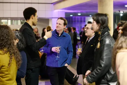 Group of people standing in a circle in the List Center atrium. The person on the far left is speaking and everyone else is looking at them while listening. The people are holding wine glasses.