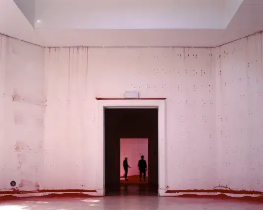 An open white-walled room with braille raised bumps across the walls. The silhouettes of two people can be seen through the dark doorway in the center of the image.