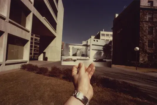 Photograph of a left hand holding up a rectangular photo of a black and white building. The photograph fits seamlessly with the street behind and surround buildings.