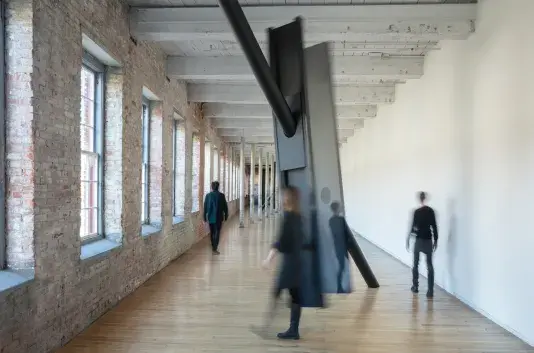 Long rectangular space with light wood floors and a series of windows on the brick wall at the left side of the frame. There are large dark metal beams suspended in the middle of the space with people walking around the sculpture.