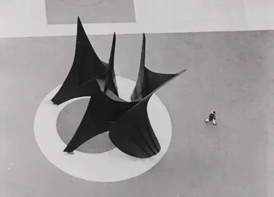 Black and white photograph of an aerial view os a large black abstract geometric sculpture. There is a person walking beside the sculpture.