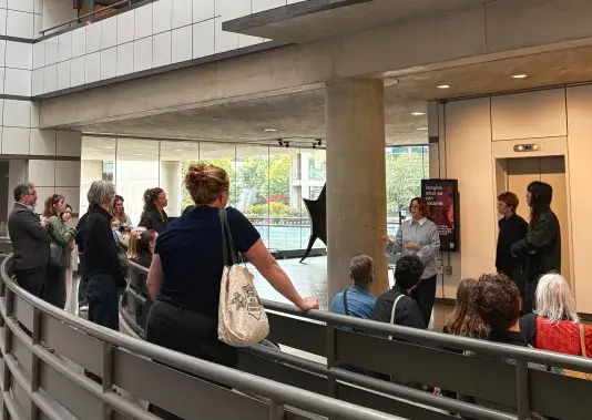 Crowd of people standing in a circle in an atrium listening to one person speak.
