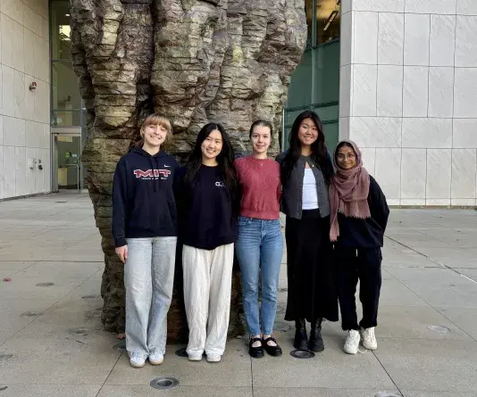 A group of five women standing side by side outside in a stone courtyard with buildings and large sculpture in the background.