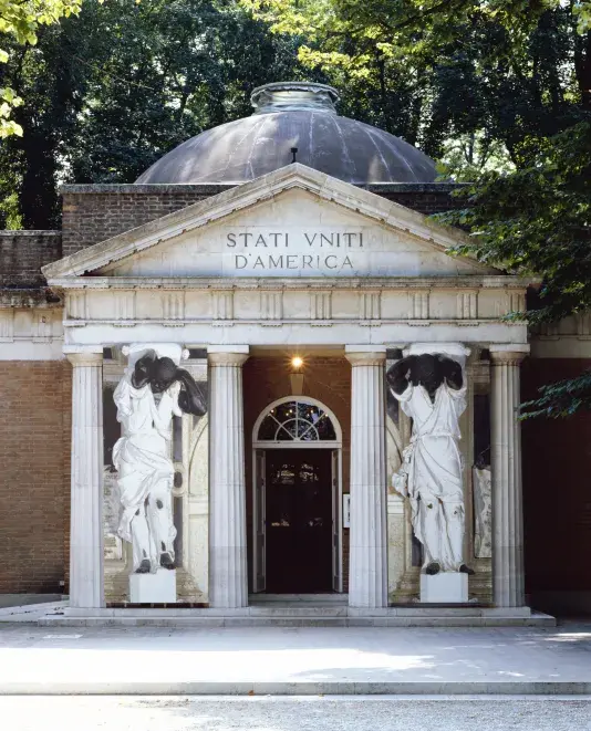 A classical stone building featuring tall columns supporting a grand entrance. There are two human figure statues on either side of the doorway. The text on the building facade reads, "STATI VNITI D'AMERICA"
