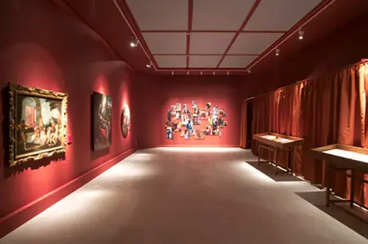 Dimly lit art gallery with deep red walls displaying framed paintings and a collage of various images. Spotlights create a focused, intimate ambiance.