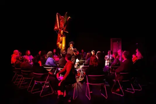 Dark room with a crowd of people highlighted against the dark background in pink light sitting and watching a woman holding a microphone in the middle of the space. There are gold metallic balloons floating to the left of the central figure.
