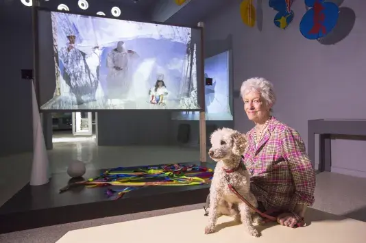 A woman with short white hair and a plaid shirt sits with a cream curly-haired dog on her lap in an art gallery. A vibrant installation featuring colorful ribbons and a large screen with abstract images is in the background.