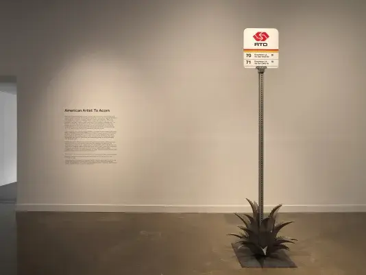 A Pasadena bus stop sign sits amidst a metal agave plant in a white gallery space with wall text saying American Artist: To Acorn on the left.