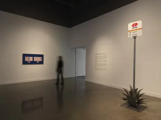 In a white gallery space, a blurred figure walks by a blue frame hung on the wall with 5 pink papers mounted inside. To the right is a Pasadena bus stop sign sitting amidst a metal agave plant.